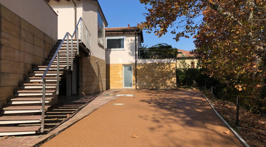 Parcheggio drenante per abitazione moderna, con scala e camminamento piastrellato a sinistra ed alberi e vegetazione a destra.