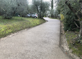 Strada sterrata pendente, in calcestruzzo drenante, con prato ed olivi a sinistra, ed altra vegetazione a destra.
