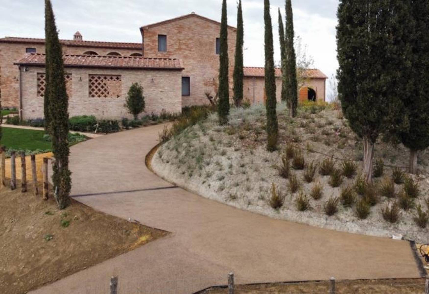 Strada ricurva e in salita in drenante chiaro, che conduce ad edifici in stile rustico, fiancheggiata da arbusti e cipressi.