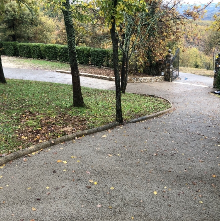 Strada privata in curva, con cancello, rivestita da pavimentazione drenante, con aiuole, foglie, alberi e siepi, in autunno.