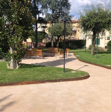Graziosa area verde con vialetti pedonali permeabili, panchine in legno ed alberi, situata in un contesto residenziale.